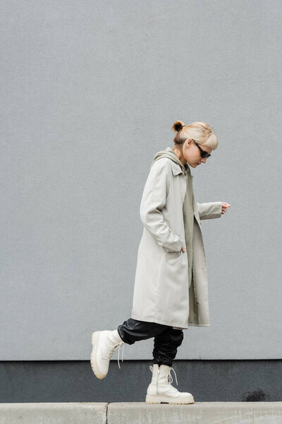full length of young woman in stylish sunglasses, long hoodie, coat, black leather pants and beige boots walking with hand in pocket near grey wall of modern building and looking down 