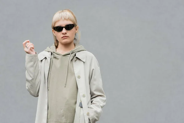 stylish and young woman with bangs and blonde hair standing in trendy sunglasses and comfortable clothes while looking at camera and posing with hand in pocket isolated on grey background in studio