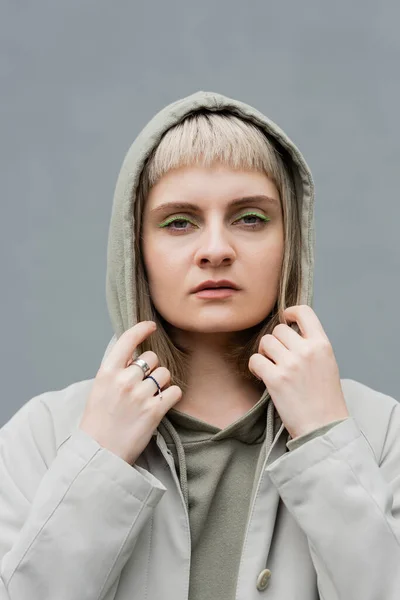 stylish and young woman with bangs and blonde hair standing with hood on head and comfortable clothes while looking at camera isolated on grey background in studio, hoodie