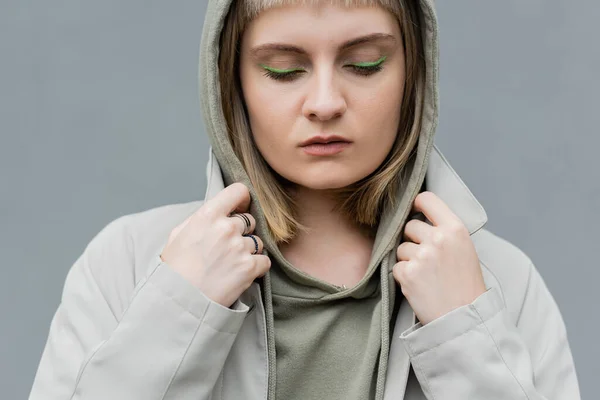 stylish and young woman with bangs, green eye shadows and blonde hair standing with hood on head and comfortable clothes while looking down isolated on grey background in studio, hoodie