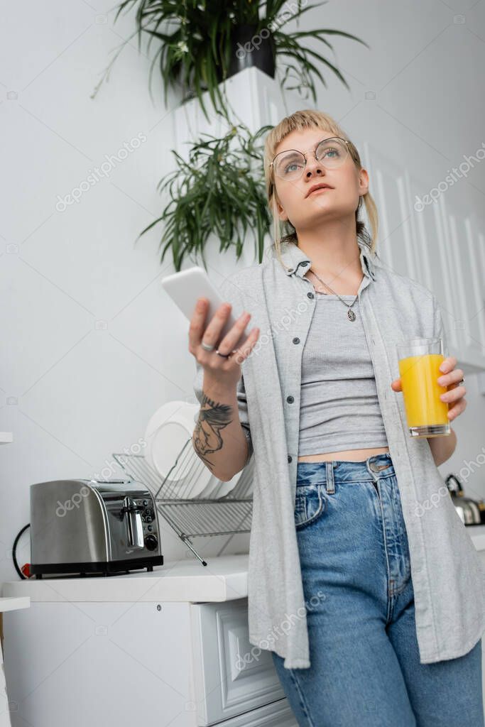 Tattooed young woman in eyeglasses, with ring on finger holding glass of orange juice and smartphone while texting and standing in blurred kitchen with toaster and indoor plants in modern apartment
