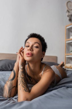 Çekici bir kadın, dövmeli seksi vücutlu, kısa esmer saçlı gri yatak örtüsüne uzanmış ve arka planda bulanık yastıkların yanındaki kameraya bakıyor.