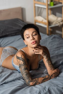 Seksi vücudu ve kısa esmer saçları olan, etkileyici dövmeli kadın, çenesine dokunuyor ve modern yatak odasındaki bulanık arka planda, bulanık arka planda, yatak odası fotoğrafçılığına bakıyor.