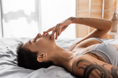 side view of young tempting woman in bra, with sexy bust and short brunette hair touching lips while laying with closed eyes on grey bedding near room divider on blurred background clipart