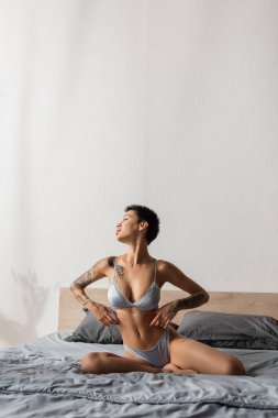 irresistible woman in silk lingerie, with short brunette hair and sexy tattooed body looking away while posing on grey bedding near pillows in modern bedroom, boudoir photography clipart