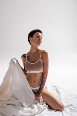 young, tattooed and passionate woman in beige lingerie, with sexy body and short brunette hair holding white bed sheet and looking away in studio on grey background, erotic photography clipart