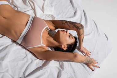 Genç, seksi ve çekici bir kadın. Kapalı gözleri, kısa esmer saçları ve dövmeli vücudu var. Stüdyodaki beyaz yatakta yatıyor ve poz veriyor.