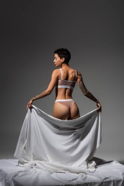 back view of stunning and sexy woman in beige lingerie, with short brunette hair and tattooed body holding white bed sheet while standing on grey background in studio, art of seduction clipart