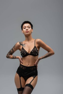 sexy tattooed woman in black glamour lingerie, bra with pearl beads, lace panties, garter belt and stockings posing with hands on hips and looking at camera on grey background clipart