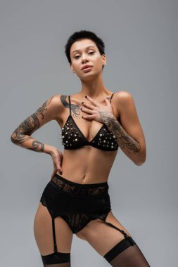Seksi dövmeli ve kısa esmer saçlı tahrik edici bir kadın, inci boncuklu sutyen, siyah külot, jartiyer kemeri ve kalçasında çorapla poz veren gri arka plandaki kameraya bakıyor.