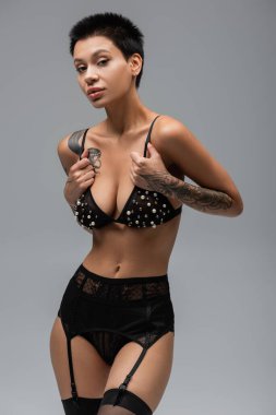 Seksi dövmeli, kısa, esmer, siyah dantelli külot, jartiyer kemeri ve çoraplı, gri arkaplanda inci boncuklu sütyen takan çekici bir kadın.