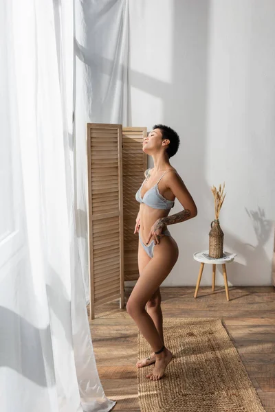 full length of stunning woman with sexy tattooed body enjoying natural light while standing on wicker rug near white curtain, room divider and bedside table with vase and spikelets in modern bedroom
