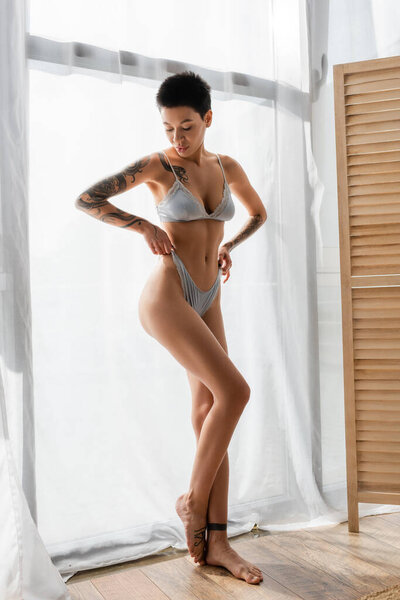 full length of young and intriguing woman with short brunette hair and sexy tattooed body pulling grey silk panties while standing near white curtain and room divider in modern bedroom