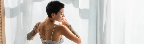 stock image back view of young, sexy and passionate woman with tattooed body and short brunette hair standing in bra near white curtain in bedroom at home, erotic photography, banner