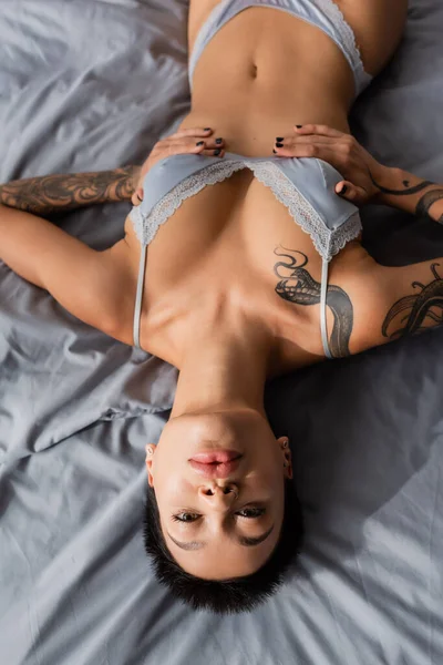 stock image top view of stunning and intriguing woman in grey silk bra, with sexy bust and short brunette hair touching slender tattooed body and looking at camera on grey bedding at home
