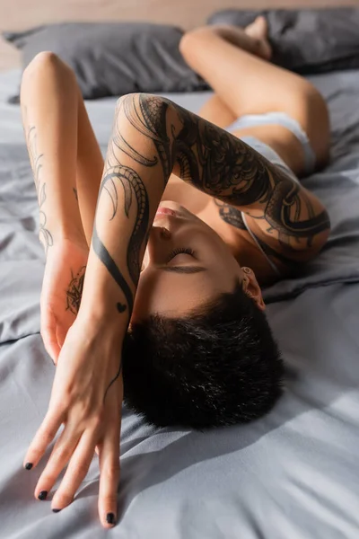young and passionate woman with closed eyes and sexy tattooed body posing in lingerie on grey bedding near pillows on blurred background in modern bedroom at home