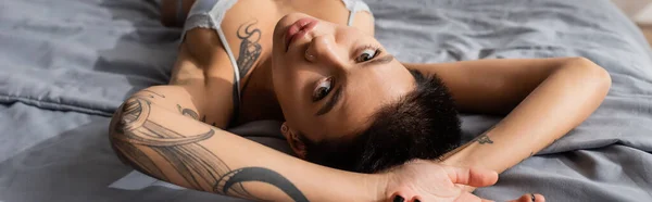 stunning woman in bra, with short brunette hair and sexy tattooed body looking at camera while laying on grey bedding in modern bedroom at home, banner