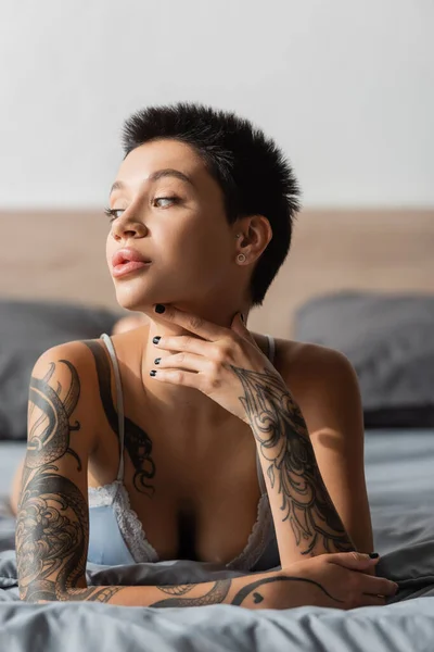 stunning woman with short brunette hair, tattooed body and sexy breast holding hand near neck while laying in bra on grey bedding and looking away on blurred background, boudoir photography