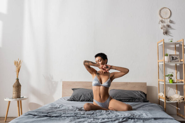 young seductive woman in silk lingerie, with sexy tattooed body and short brunette hair posing on grey bedding near rack, dream catcher, bedside table and wicker vase with spikelets in bedroom