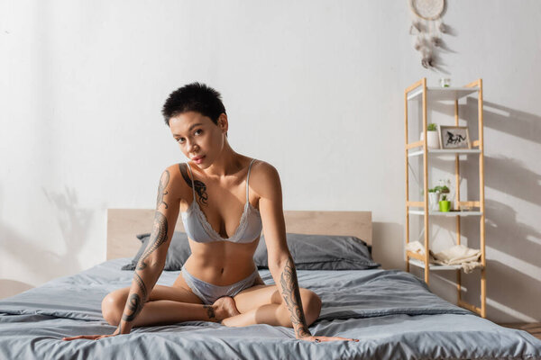 young charming woman in silk lingerie, with tattooed body and short brunette hair sitting in provocative pose and looking at camera on grey bedding near pillows, dream catcher and rack in bedroom