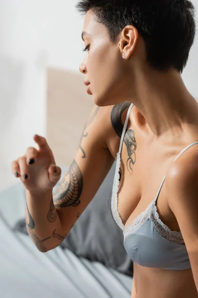 年轻的 纹身的 迷人的女人 穿着丝绸胸罩 性感的胸部 短短的黑发 在现代卧房 闺房摄影的模糊背景下做手势 — 图库照片