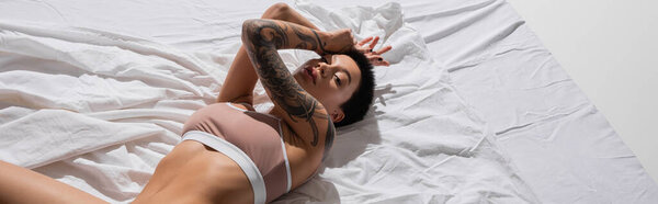 high angle view of flirtatious woman in beige lingerie, with sexy tattooed body and short brunette hair laying on white bedding and looking at camera, erotic photography, banner