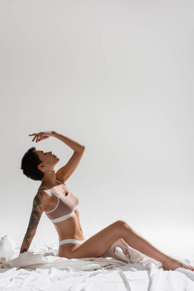 side view of young, sexy and charming woman in beige lingerie, with short brunette hair and tattooed body sitting with hand above head on white bedding and grey background, erotic photography