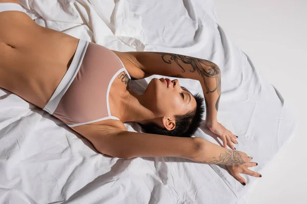 top view of young, sexy and desirable woman with closed eyes, short brunette hair and tattooed body laying and posing on white bedding in studio, erotic photography, art of seduction