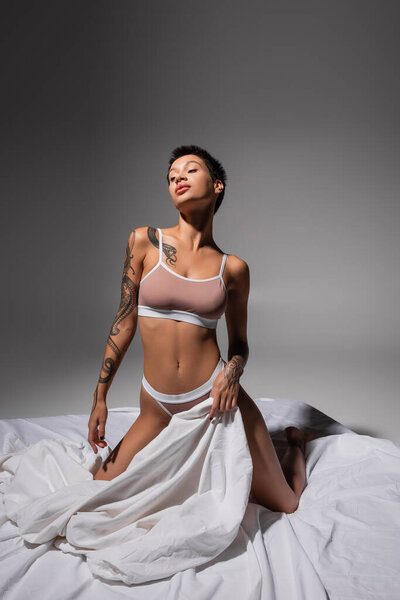 young and passionate tattooed woman in beige lingerie, with slender and sexy tattooed body and short brunette hair posing on white bedding and grey background in studio, art of seduction