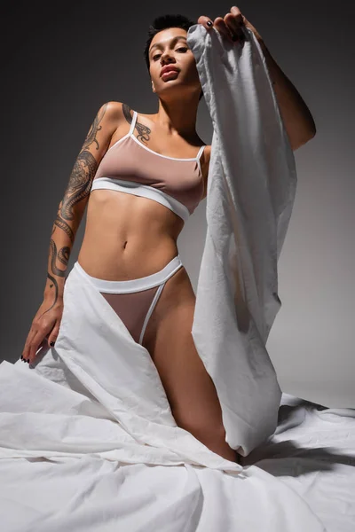 low angle view of provocative woman with short brunette hair and sexy tattooed body holding white bed sheet and posing in beige lingerie on grey background in studio, erotic photography