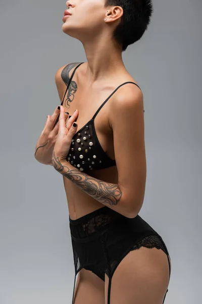 stock image cropped view of young, graceful and tattooed woman in black bra with pearl beads, lace panties and garter belt holding hands near chest on grey background in studio, art of seduction