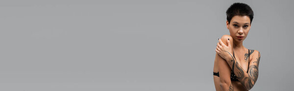 молодая, стильная и провокационная женщина в черном лифчике, с татуированным телом и короткими волосами брюнетки, обнимающая себя и смотрящая на камеру на сером фоне, эротическая фотография, баннер