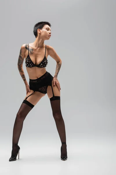 full length of expressive woman with sexy tattooed body wearing black bra with pearl beads, lace panties, garter belt, stockings with high heels and posing with hands on hips on grey background