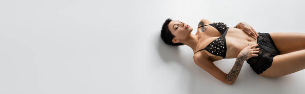 высокий угол обзора молодой заманчивой женщины с закрытыми глазами, короткие волосы брюнетки и сексуальное татуированное тело, лежащее в бюстгальтере с жемчужными бусами и черными кружевными трусиками на сером фоне, баннер