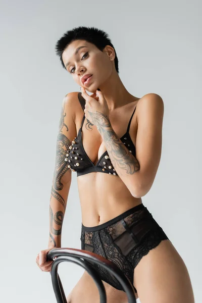sexy and flirtatious woman with tattooed body and short brunette hair, wearing black bra with pearl beads and lace panties while looking at camera near chair on grey background