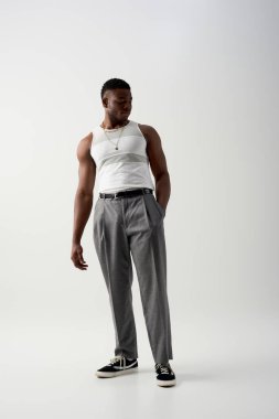 Kolsuz tişörtlü ve pantolonlu, gri arka planda duran, modern çekimde şık giyinen, uzun boylu Afro-Amerikan bir adam.