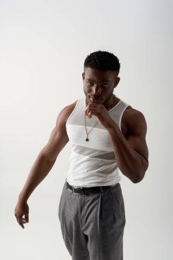 Askılı tişörtlü, pantolonlu, kendine güveni tam genç Afroamerikan bir adam kameraya bakarken gri, çağdaş çekimlerde şık kıyafetler, moda tasarımı... 