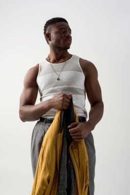 Askılı tişört giyen kaslı Afroamerikalı genç bir adam ve moda bombacısı ceketi tutan pantolon gri renkli, çağdaş çekimlerde şık giyinen, moda demecinde 