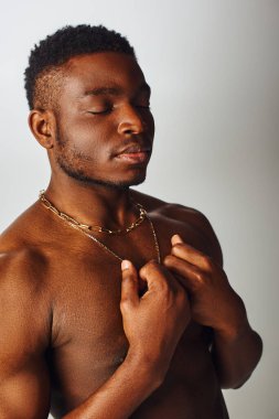Kaslı ve üstsüz Afro-Amerikan erkeğinin portresi. Gözleri kapalı. Gri, özgüvenli ve modern pozlarda izole edilmiş kolyelere dokunuyor.