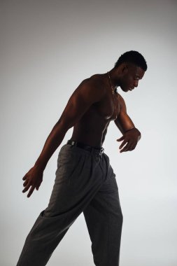 Side view of muscular african american model in gray pants standing in shadow isolated on grey, confident and modern pose, fashion shoot, shirtless man clipart