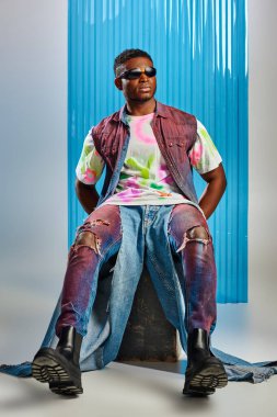 Güneş gözlüğü, kot yelek ve yırtık kot pantolon giymiş, arka planda mavi polikarbonat çarşaf, moda çekimi ve sürdürülebilir moda ile taşın üzerinde oturan Afro-Amerikan modelleri. 