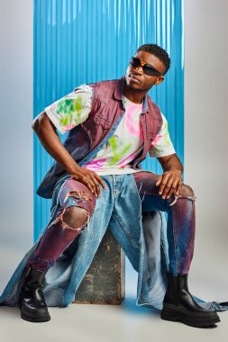 Young african american man with trendy hairstyle in colorful t-shirt and denim vest sitting on stone on grey with blue polycarbonate sheet at background, fashion shoot, DIY clothing clipart