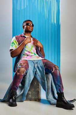 Güneş gözlüklü, renkli kot yelekli ve tişörtlü modaya uygun Afro-Amerikan modeli taşın üstünde gri üzerine oturmuş arka planda mavi polikarbonat çarşafı, moda çekimi, DIY giysi