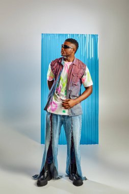 Full length of trendy african american model in colorful t-shirt, sunglasses and ripped jeans standing on grey with blue polycarbonate sheet at background, fashion shoot, sustainable lifestyle clipart