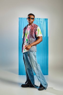 Full length of stylish young afroamerican model in sunglasses, ripped jeans and denim vest standing on grey with blue polycarbonate sheet at background, fashion shoot, DIY clothing clipart