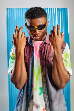 Portrait of stylish young afroamerican model in sunglasses, colorful denim vest and t-shirt posing and standing on grey with blue polycarbonate sheet at background, sustainable fashion, DIY clothing clipart