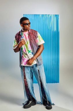 Güneş gözlüklü, yırtık kot pantolonlu ve renkli yelekli modaya uygun arka planda mavi polikarbonat çarşaflı gri üzerine poz veren Afroamerikan modelleri.