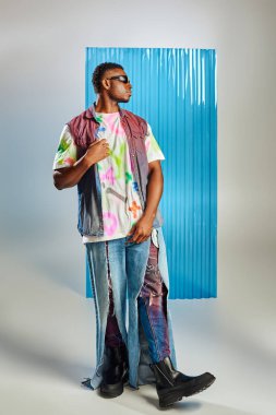 Güneş gözlüklü şık Afroamerikan erkeği, renkli kot yelek ve arka planda mavi polikarbonat çarşaflı yırtık kot pantolon, sürdürülebilir moda, DIY giysi
