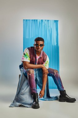 Güneş gözlüklü Afroamerikan modeli, renkli kot yelek ve çağdaş yırtık kot pantolon taşın üzerinde duruyor arka planda mavi polikarbonat örtüsü, sürdürülebilir moda, DIY giysi