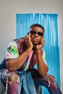 Portrait of young afroamerican model in sunglasses and colorful denim vest and ripped jeans sitting on grey with blue polycarbonate sheet at background, sustainable fashion, DIY clothing clipart